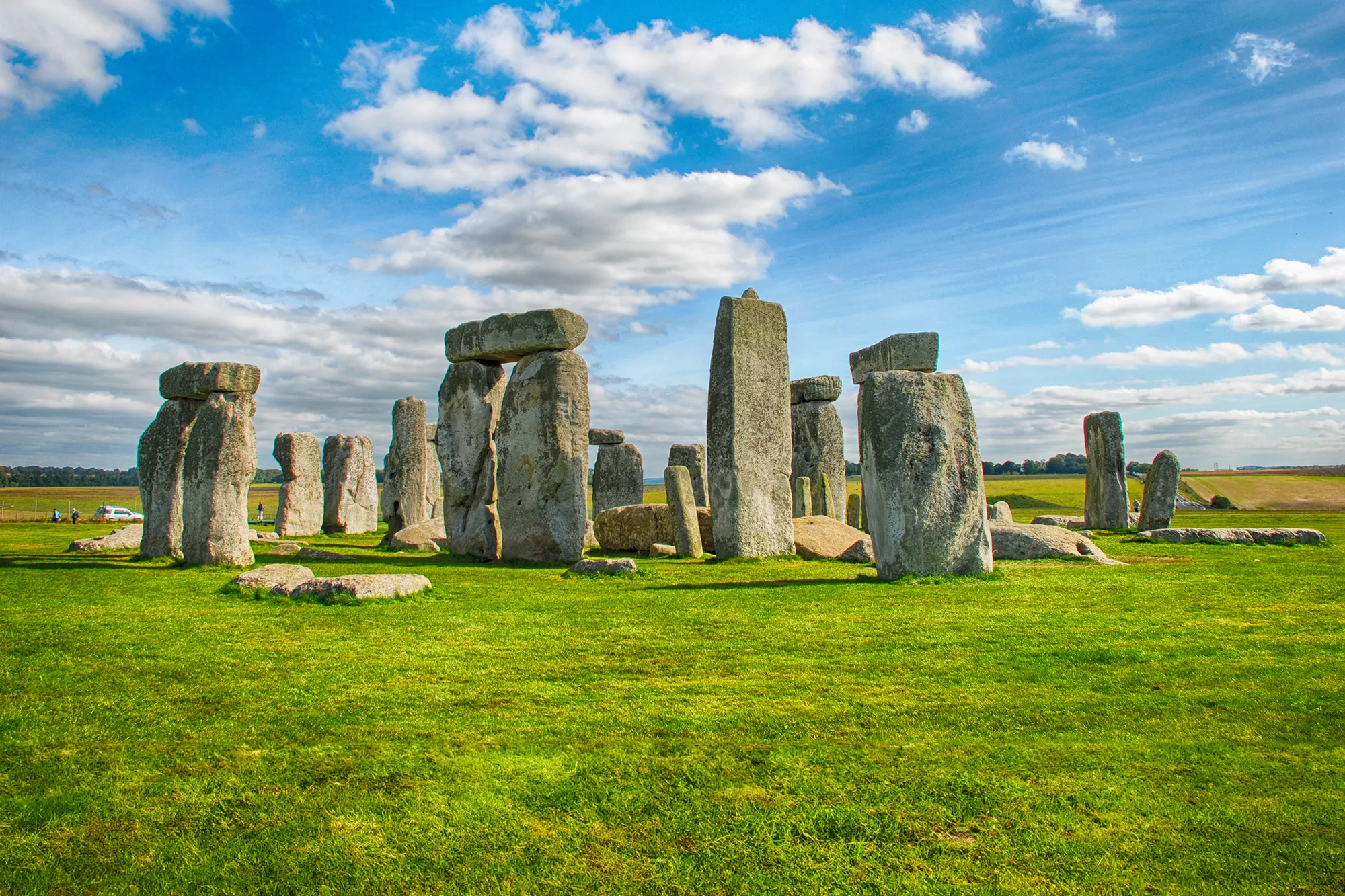 A bright, clear image of Stonehenge, an ancient prehistoric monument consisting of a circular arrangement of massive standing stones. The stones are set against a vibrant green grass field with a blue sky and scattered white clouds above.