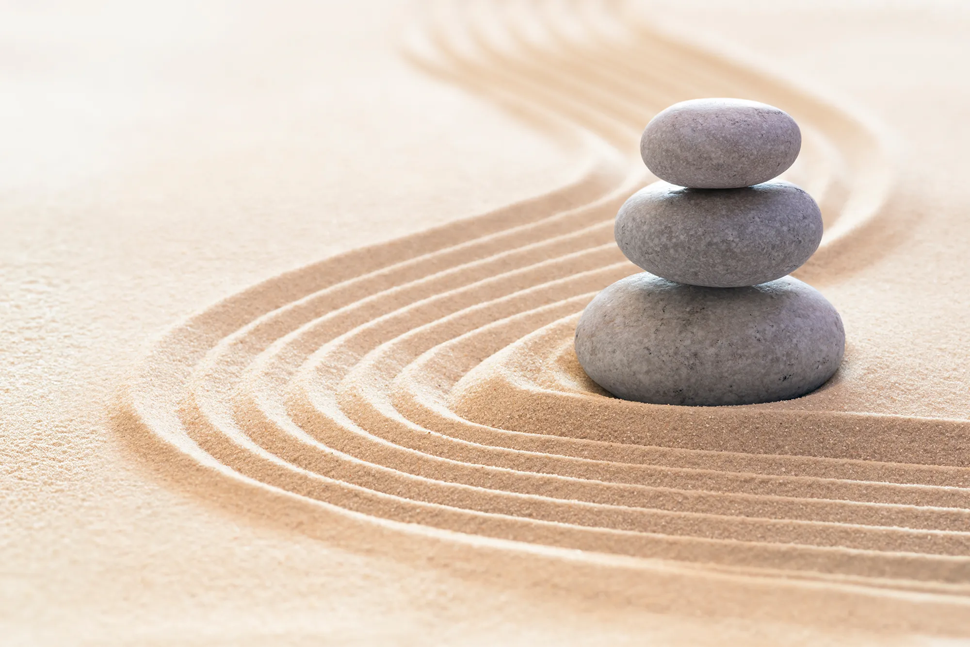 Stack of three smooth gray stones balanced on raked sand with curved lines in a Zen garden, representing peace, mindfulness, and balance.
