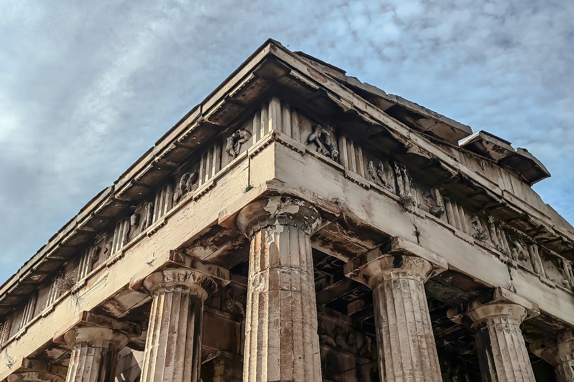 Low-angle view of an ancient Greek temple with detailed stone carvings and weathered columns against a partly cloudy sky, representing history, philosophy, and classical studies.