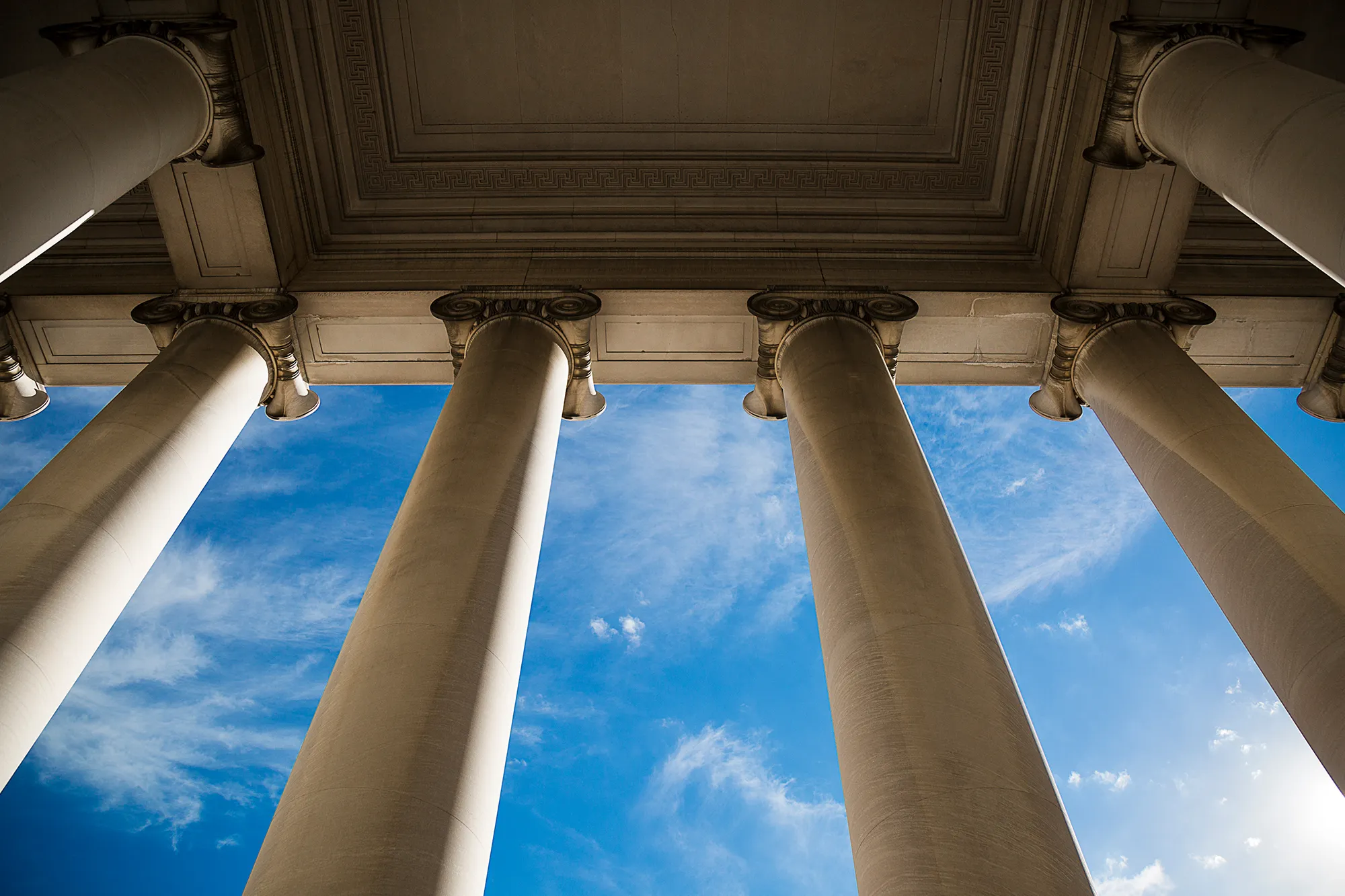 Upward view of classical stone columns and architectural detailing against a blue sky, symbolizing the foundations of liberal arts and sciences education.