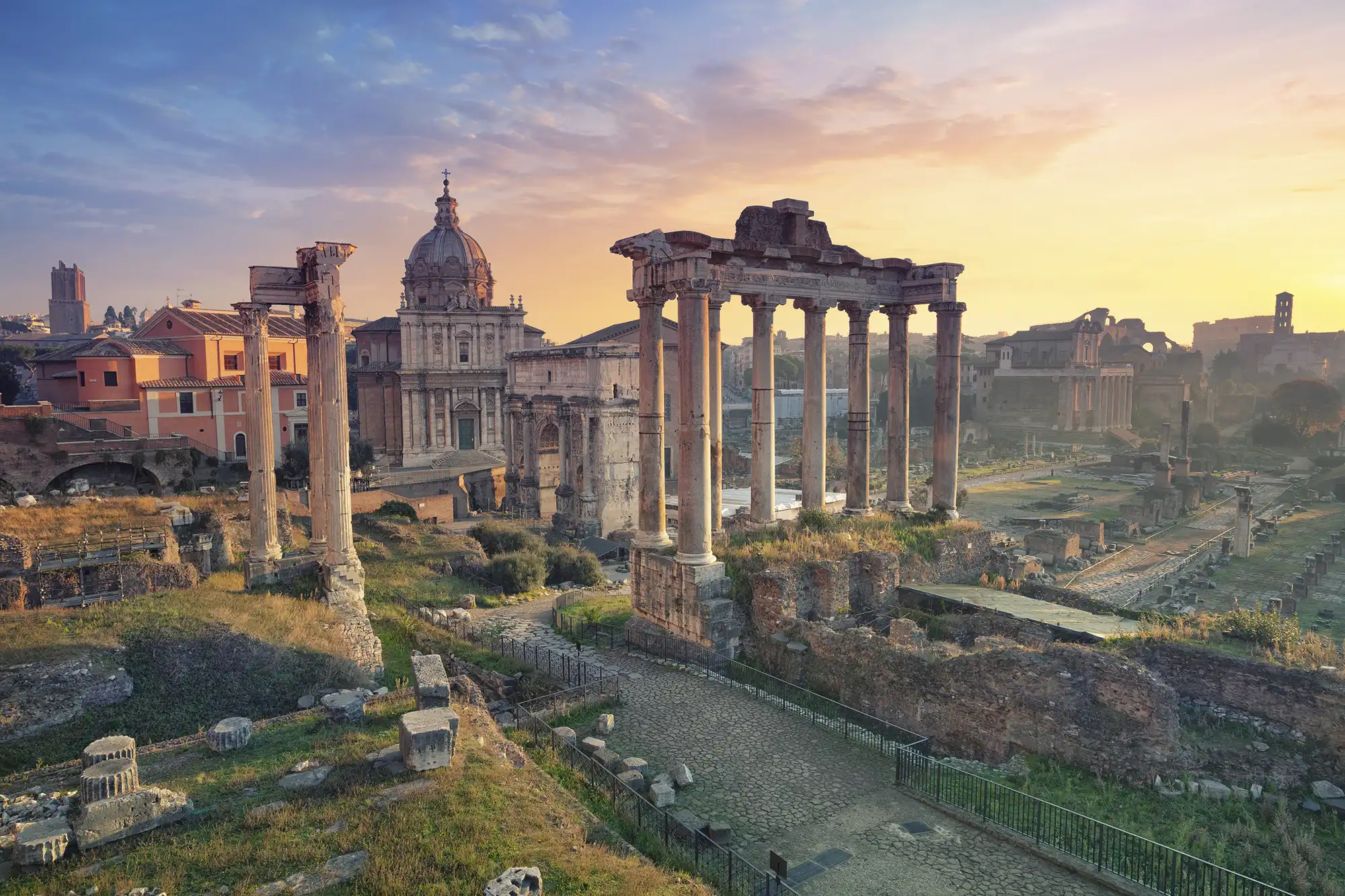 A view of the Roman Forum at sunset, featuring the remaining Corinthian columns of the Temple of Saturn and the Arch of Septimius Severus against a soft pink and orange sky.