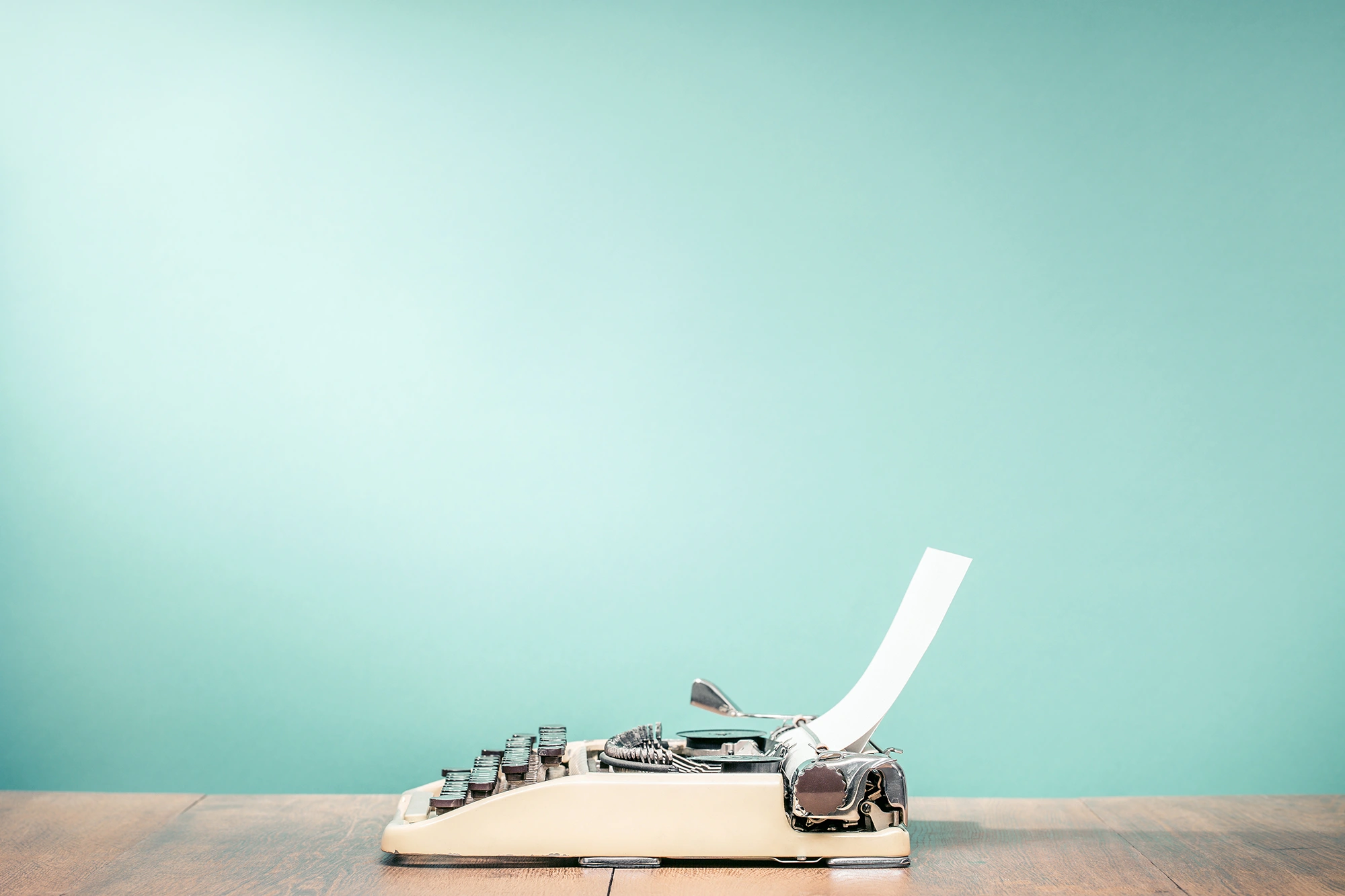 A vintage cream-colored typewriter sits on a wooden desk with a blank sheet of white paper inserted. The background is a solid, light teal color with a soft vignette effect.