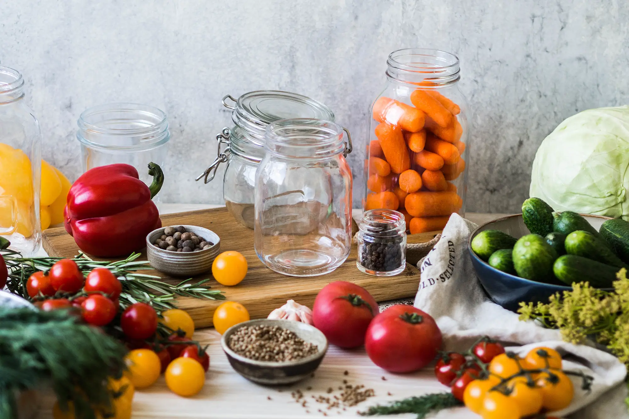 A collection of fresh produce for pickling, featuring glass jars, baby carrots, cherry tomatoes, cucumbers, and a red bell pepper. Small bowls of spices and herbs sit on a wooden cutting board.