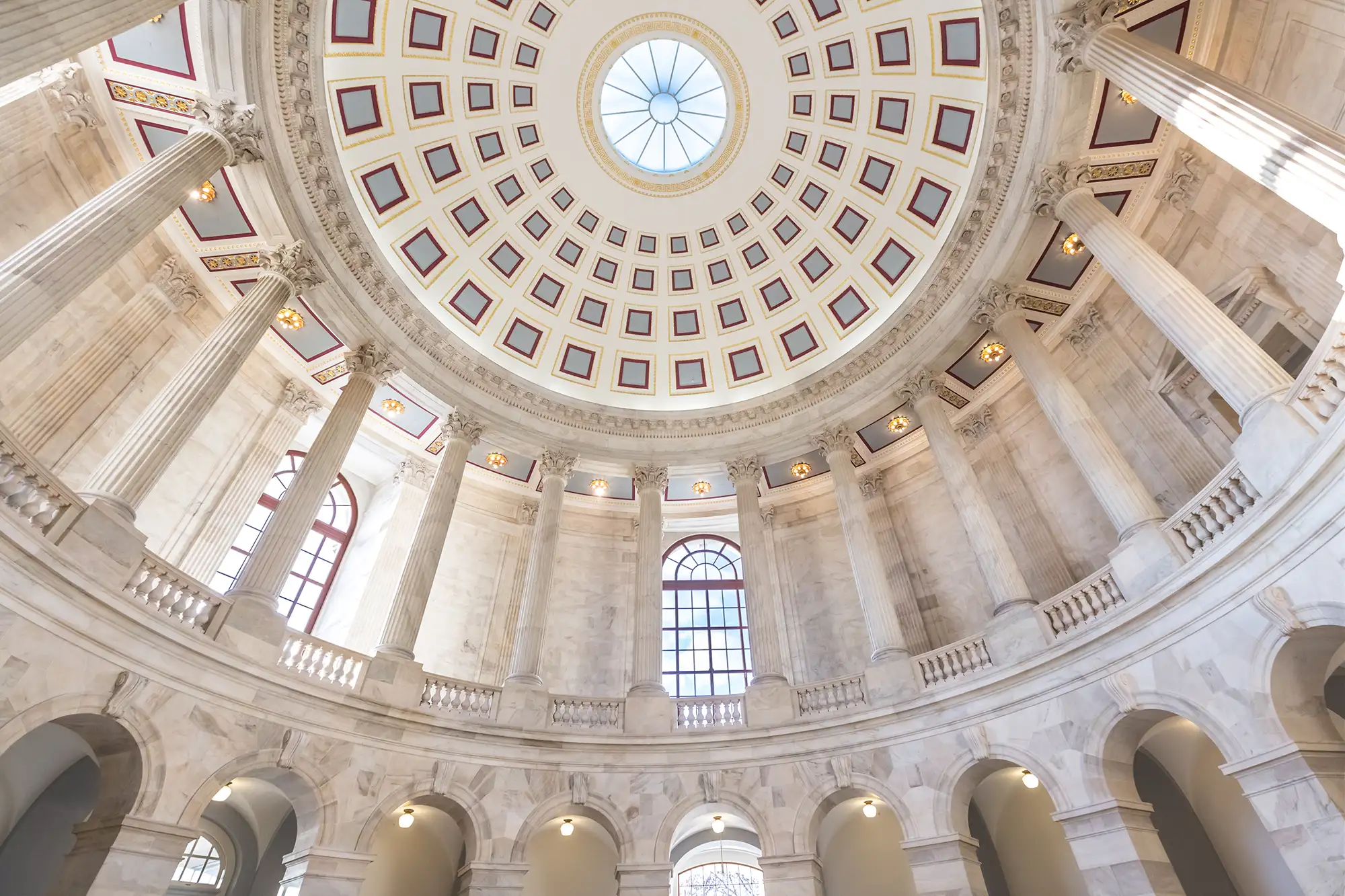 A low-angle view of a grand marble rotunda featuring a high domed ceiling with a central skylight, ornate geometric patterns, and massive fluted columns surrounding an upper gallery.