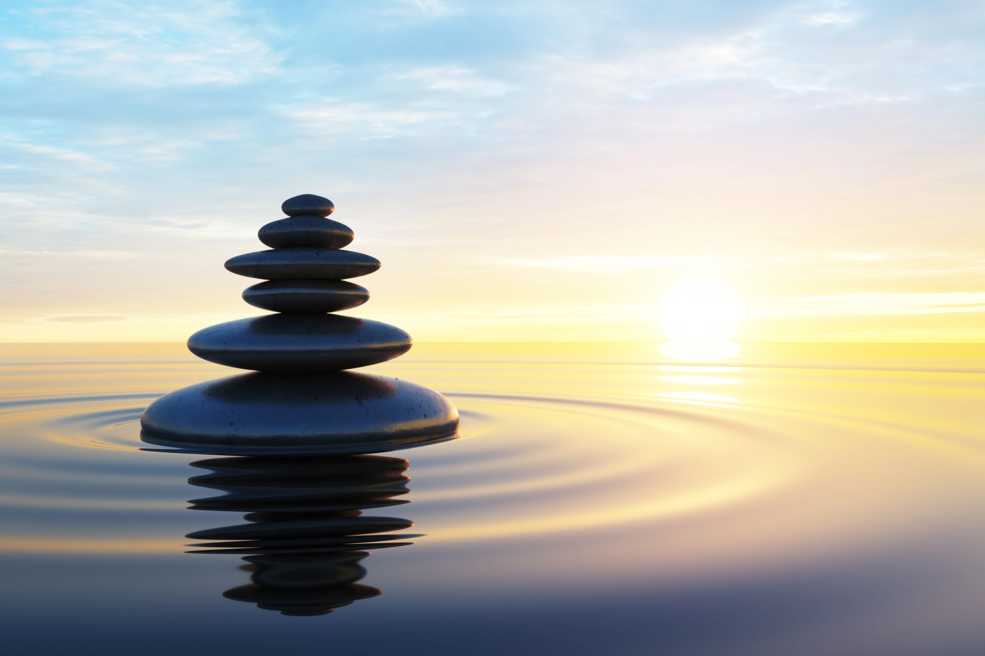 A stack of smooth, balanced stones sits in the center of calm water, creating gentle ripples. A bright sun sets on the horizon, casting a warm yellow glow over the peaceful blue sky.